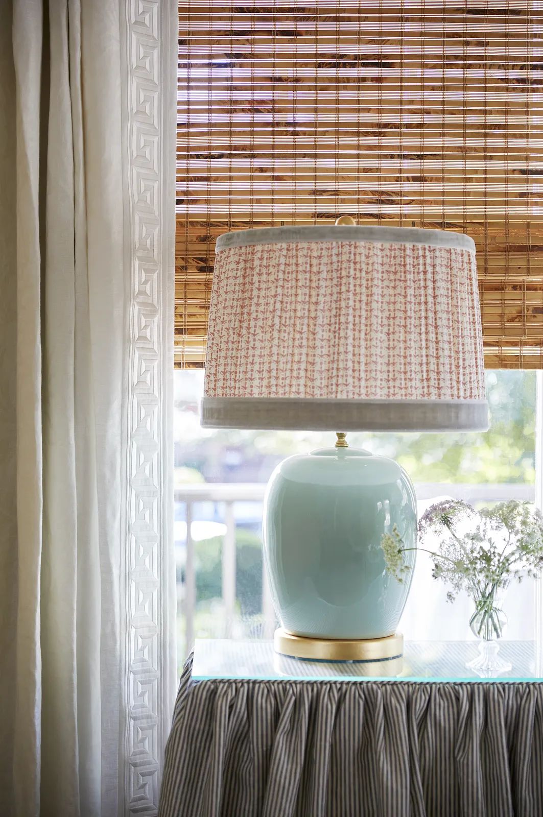 Tabletop with blue lamp, patterned shade, vase of flowers, striped skirt, and window treatments.