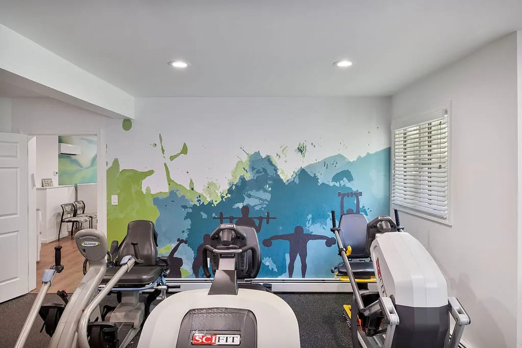 Workout space with exercise machines, colorful mural, window blinds, and black rubber flooring.