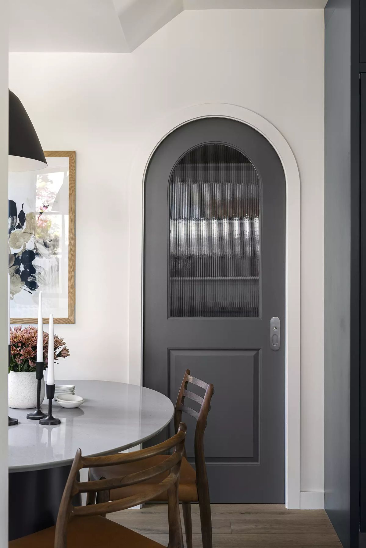 Dining room with round white table, wooden chairs, dark arched door, floral artwork, and candles.