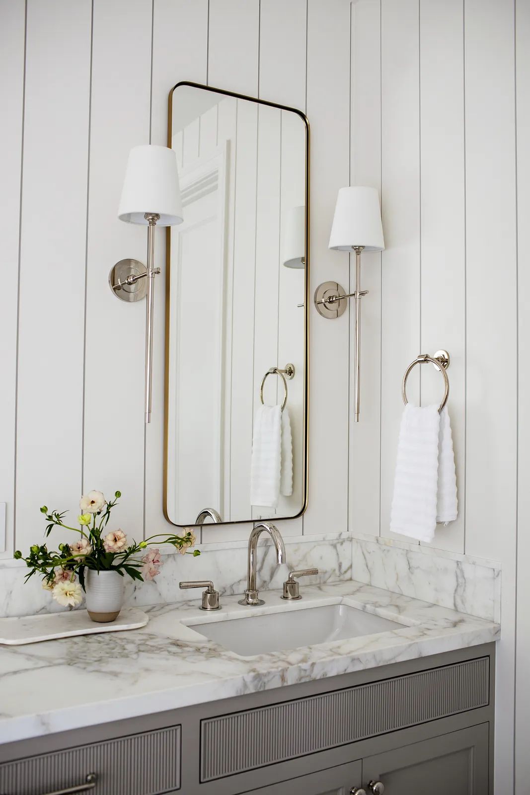 Bathroom with marble countertop, sink, white paneling, large mirror, wall sconces, and flowers.