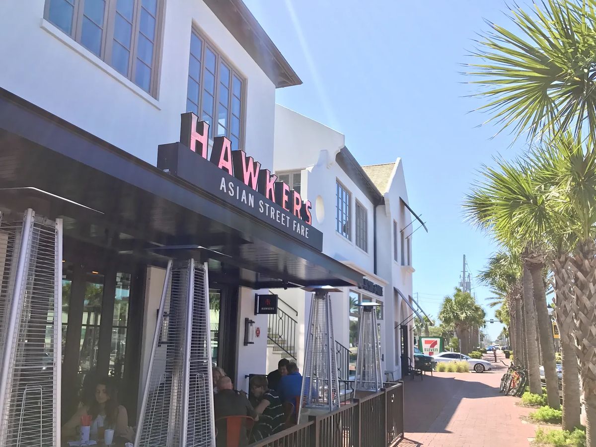 Exterior view of restaurant with black awning, white walls, outdoor seating, palm trees, and nearby 7-Eleven.