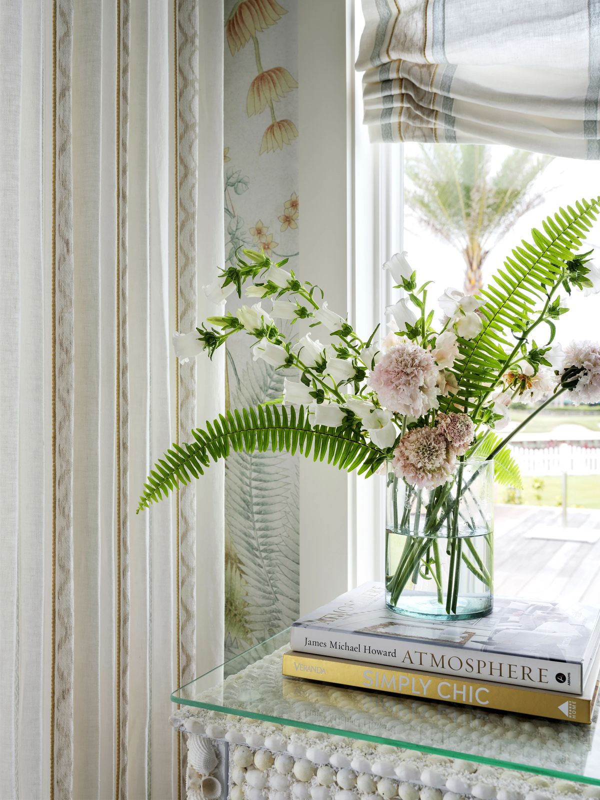 Arrangement with glass vase of flowers on decorative table, sheer curtains, and botanical patterned wall.