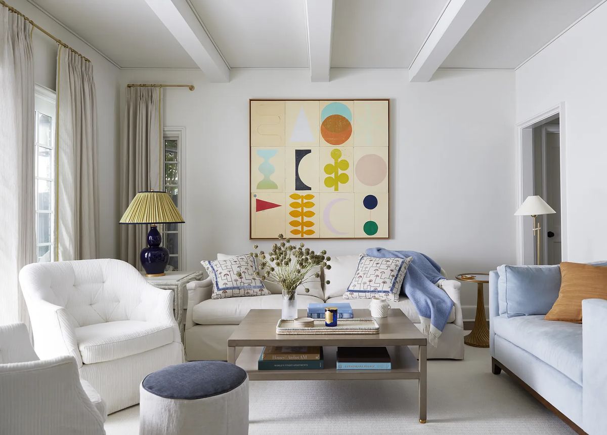 Living room with white sofas, blue throw blanket, round stool, wooden coffee table, and geometric artwork.