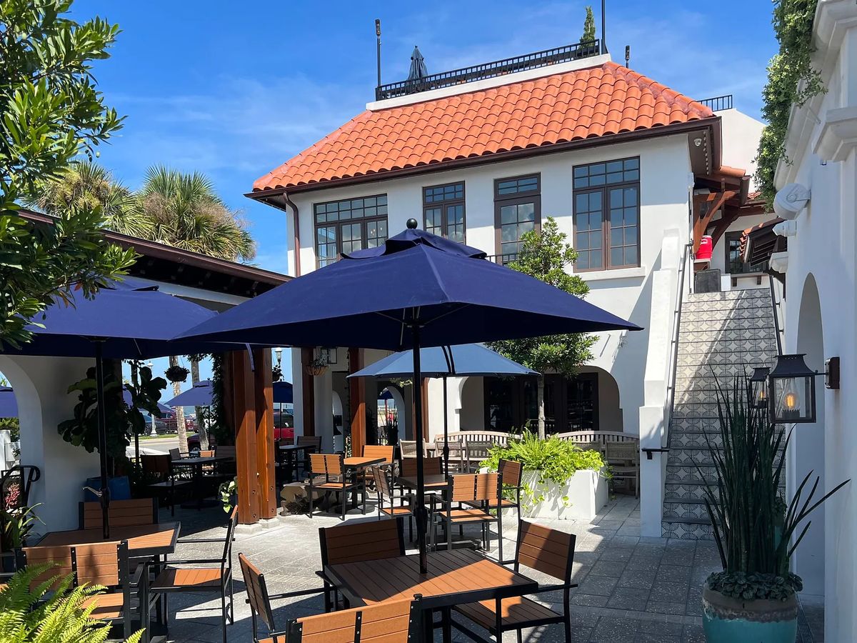 Outdoor patio with wooden tables, blue umbrellas, potted plants, and a two-story building in the background