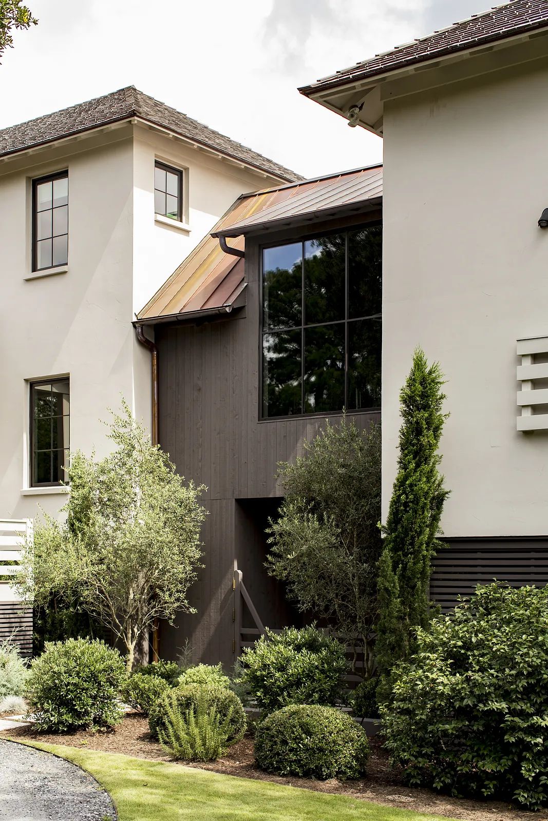 Exterior front view of a house with distinct building sections, large window, shrubs, and lawn.