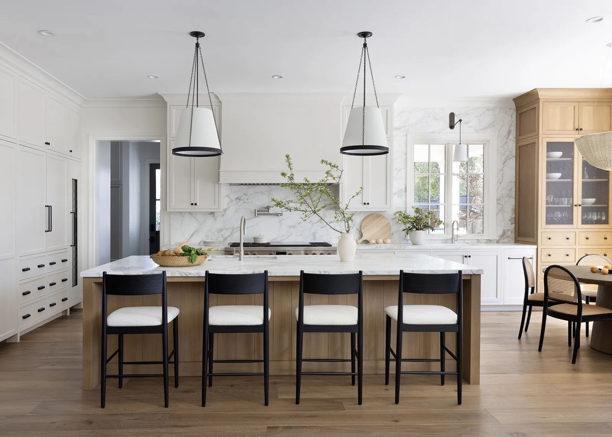 Kitchen with marble island, black bar stools, white cabinetry, two pendant lights, and large windows for natural light.