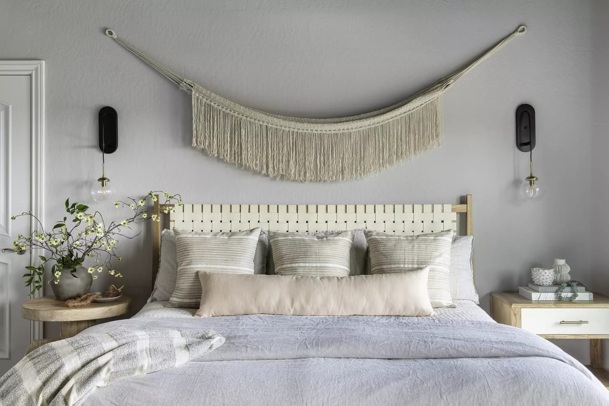 Bedroom with wooden headboard, multiple pillows, light blue quilt, nightstands, plant, and wall sconces.