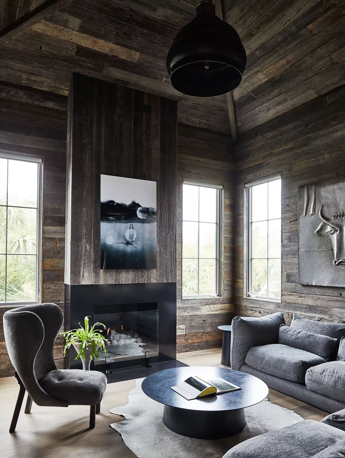 Living room with gray sofa, accent chair, round black coffee table, and large windows in wood-paneled space.