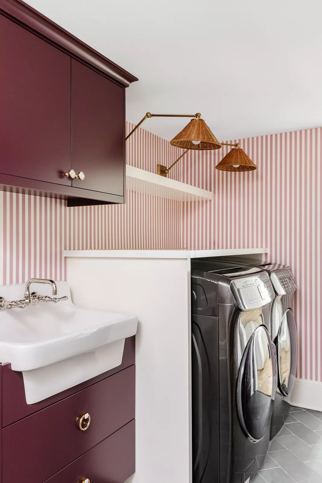 Laundry room with stacked washers, white sink, maroon cabinets, and pink striped wallpaper.