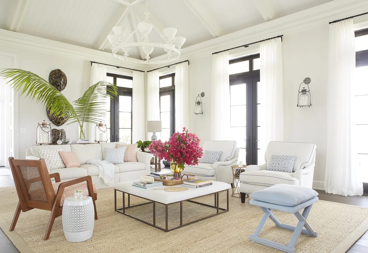 Living room with white sofa, armchairs, coffee table, area rug, plant, floral centerpiece, and tall windows with curtains