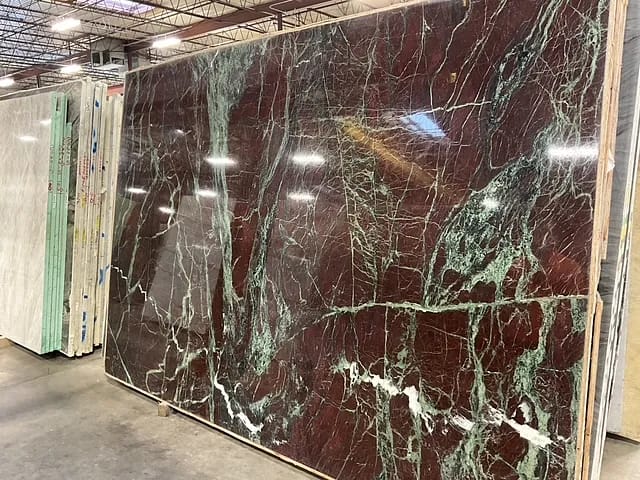 Warehouse displaying large red and green marble slabs with overhead fluorescent lighting.