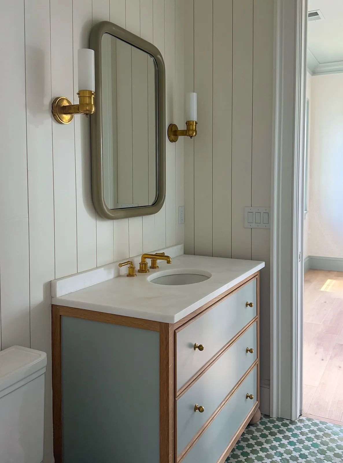 Bathroom with wooden vanity, round mirror, brass faucet, wall sconces, and patterned floor tiles.