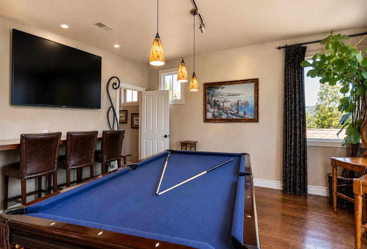 Game room with blue pool table, wall-mounted TV, bar stools, and a large indoor plant.