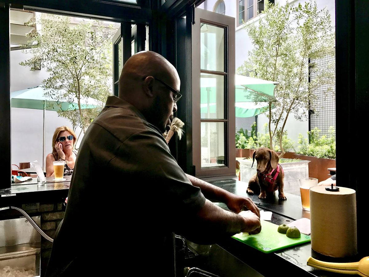 Outdoor setting with a man cutting limes, a dog on the ledge, and seated people with green umbrellas.