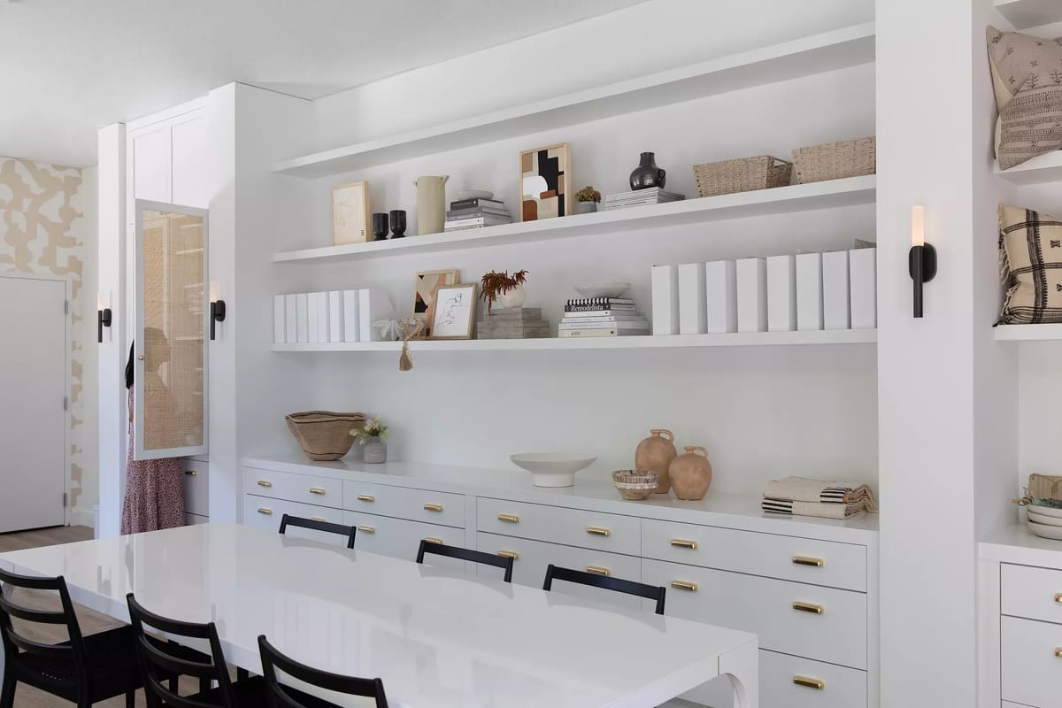 Dining room with white table, black chairs, built-in shelves, decorative baskets, and patterned wallpaper.