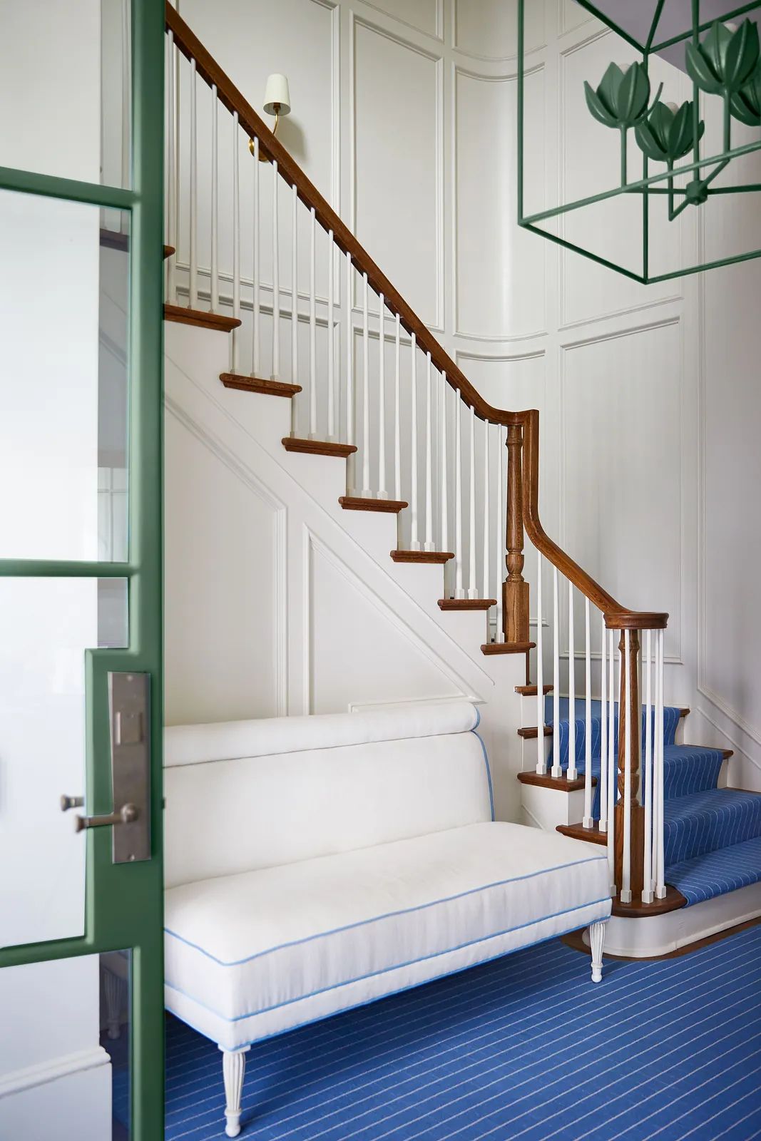 Foyer with white settee, wooden staircase, striped blue rug, and green-framed glass door.