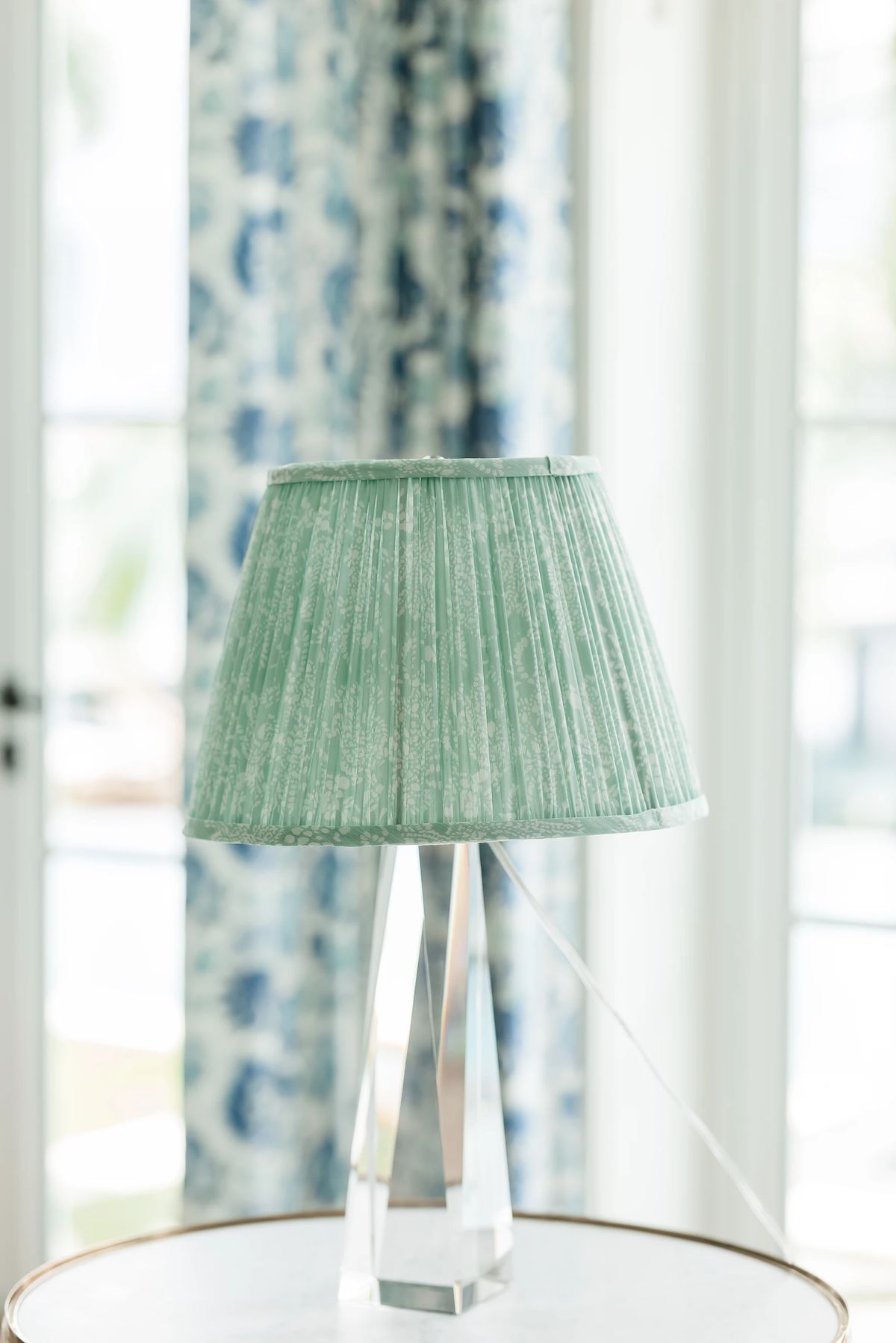 Decorative lamp with green shade on round table, floral-patterned curtains in the background.