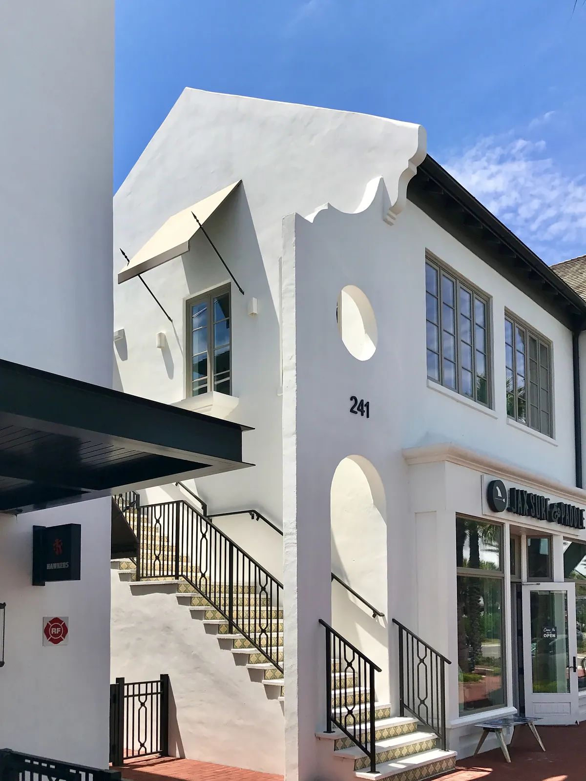 Exterior front of a white stucco building with large windows, staircase, and storefront.