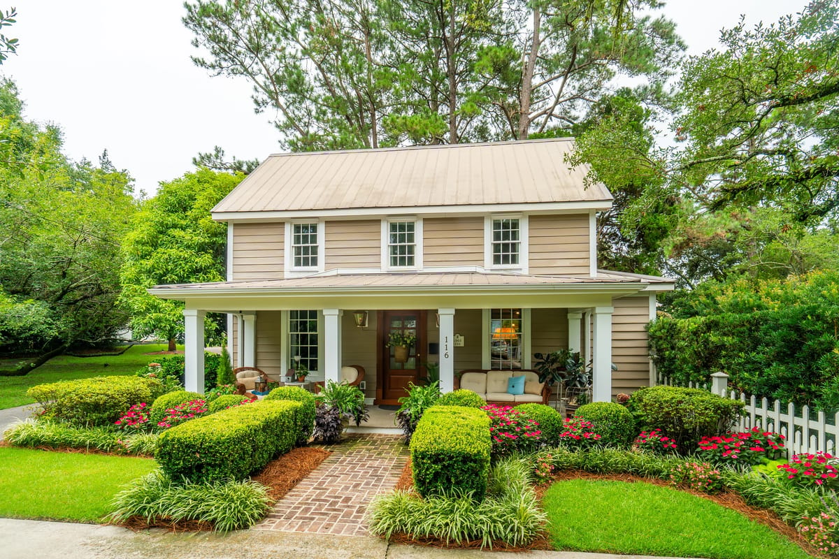 Front exterior of a house with a covered porch, wicker chairs, brick pathway, and landscaped gardens.