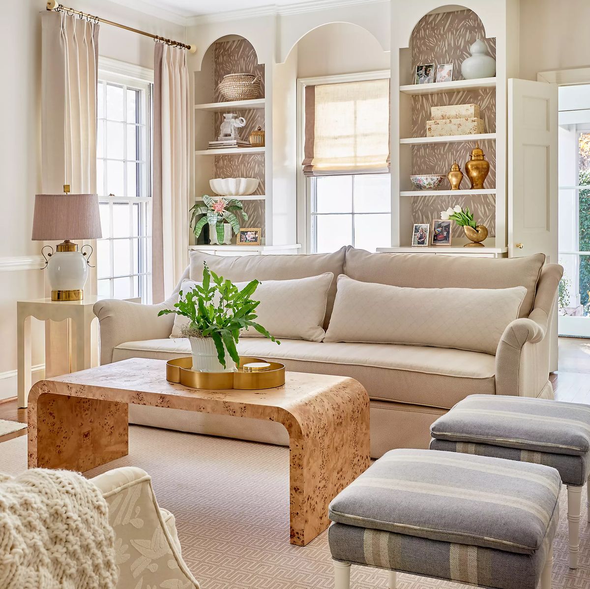 Living room with beige sofa, cork table, striped ottomans, and decorative plant by bookshelves and large windows.