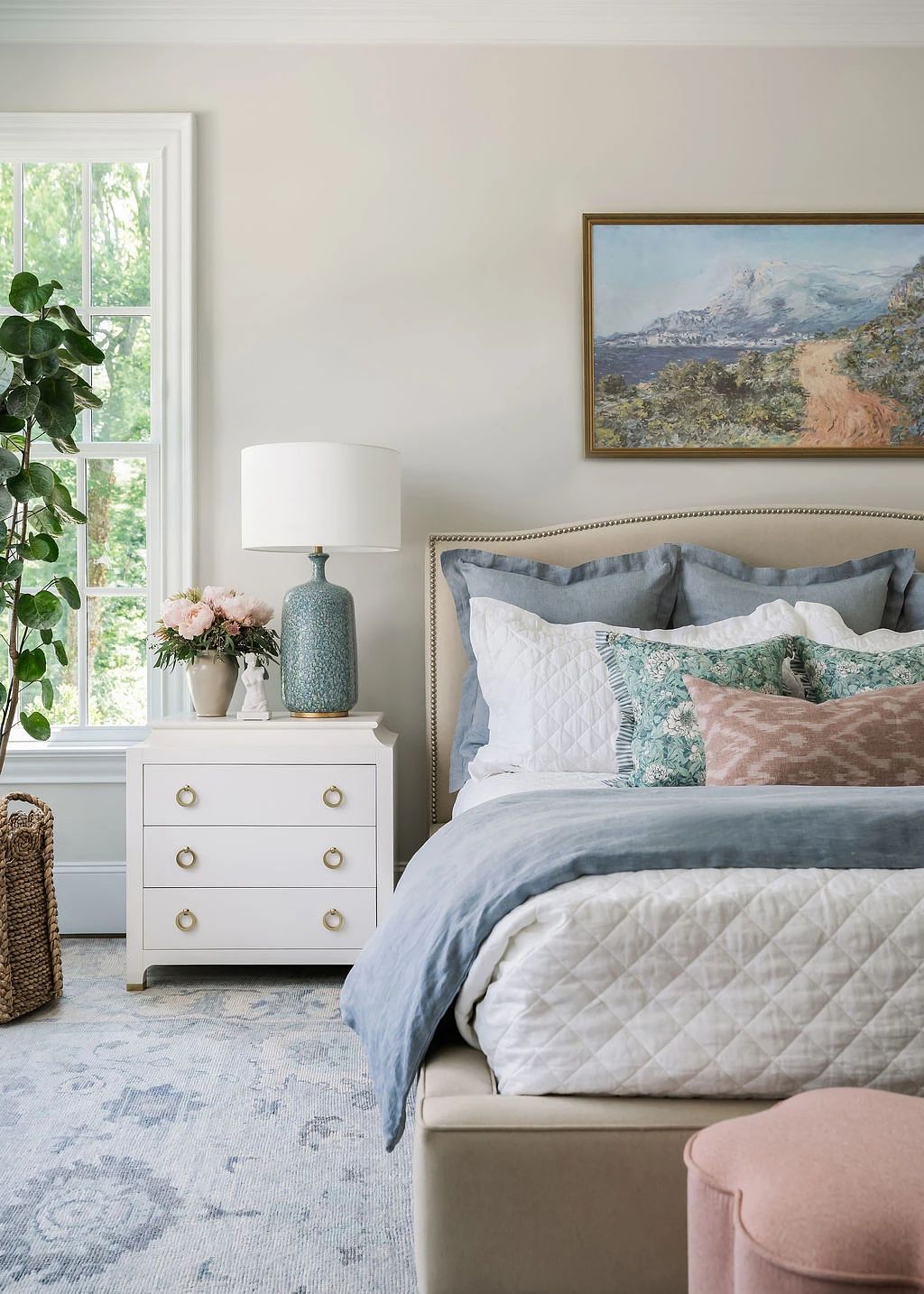 Bedroom with upholstered bed, nightstand, lamp, painting, plant, window, and pink ottoman