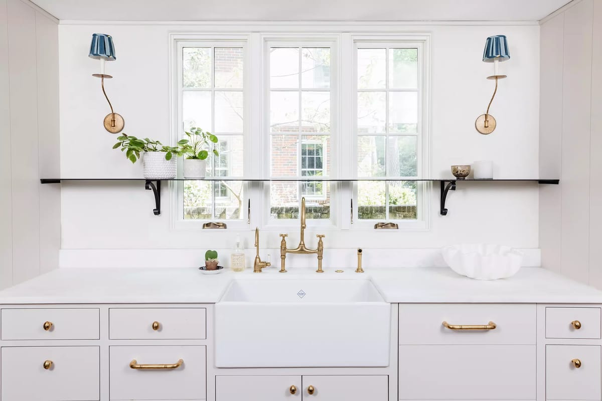 Kitchen with white farmhouse sink, gold faucet, white cabinetry, black shelf, and wall sconces near window.