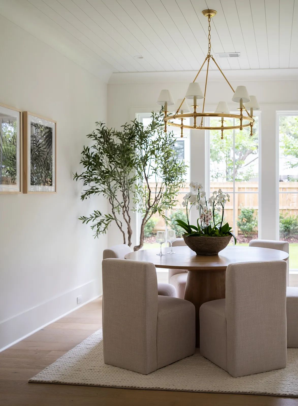 Dining room with round wooden table, upholstered chairs, potted plant, windows, artworks, and chandelier