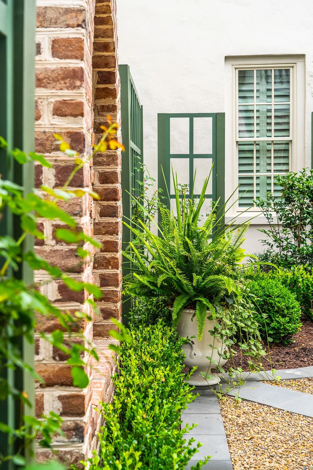 Garden with brick wall, white planter containing fern, shrubs, and stone pathways.
