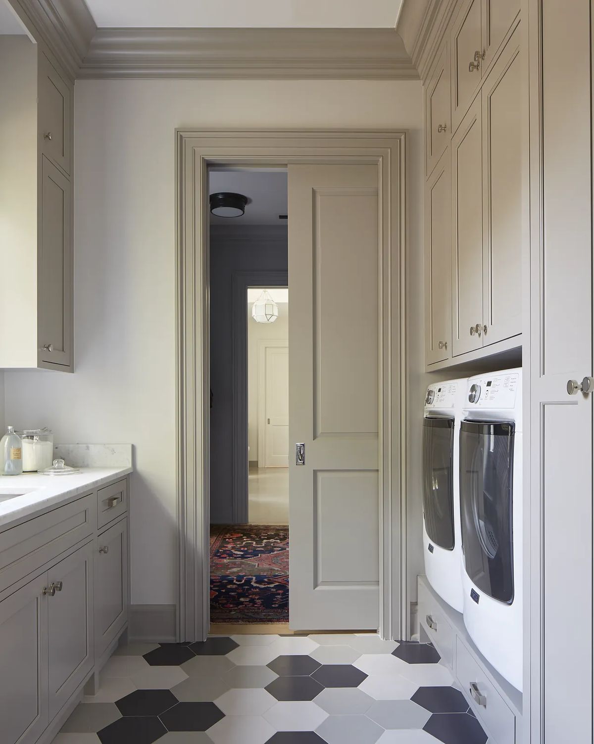 Laundry room with gray cabinetry, marble countertop, front-loading washers, and hexagonal black and white tiles.