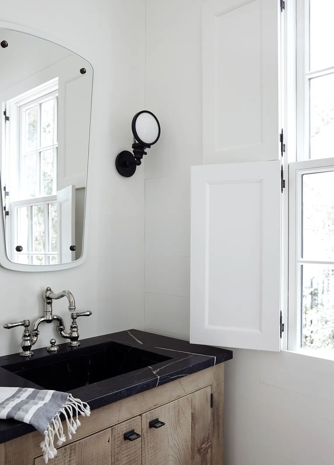 Bathroom with wooden vanity, black countertop, round mirror, and two windows with white shutters.