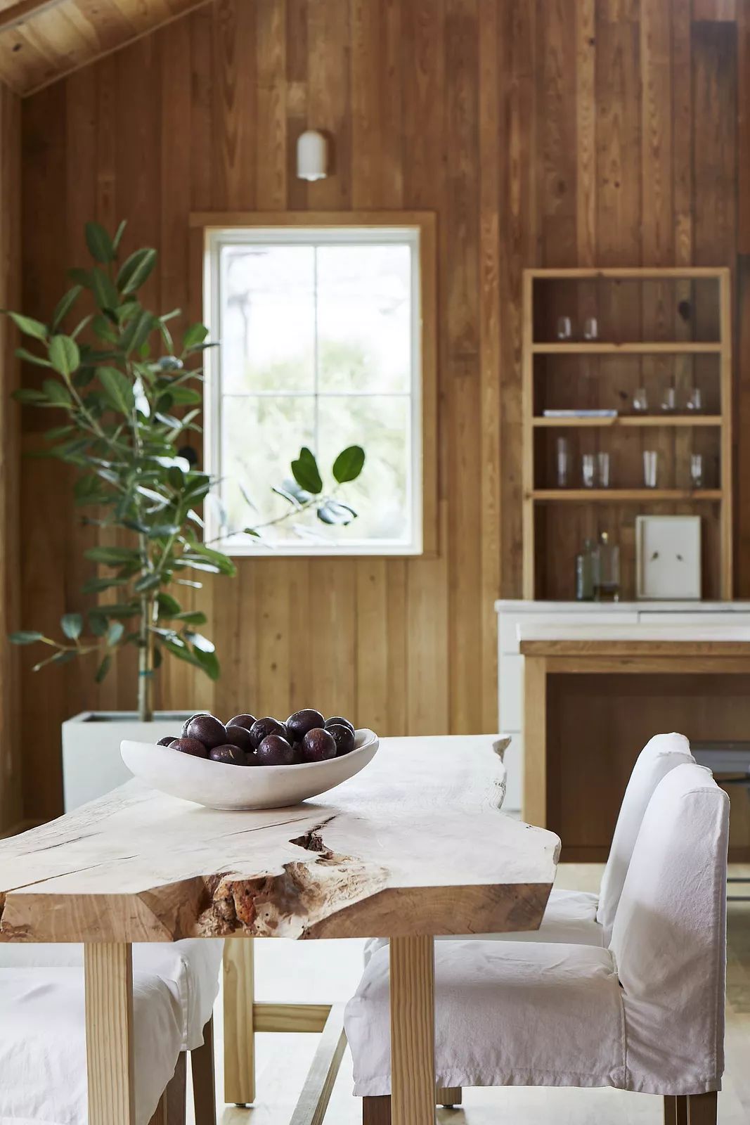 Dining room with wood table, white chairs, bowl of fruit, window, shelving unit, and potted plant