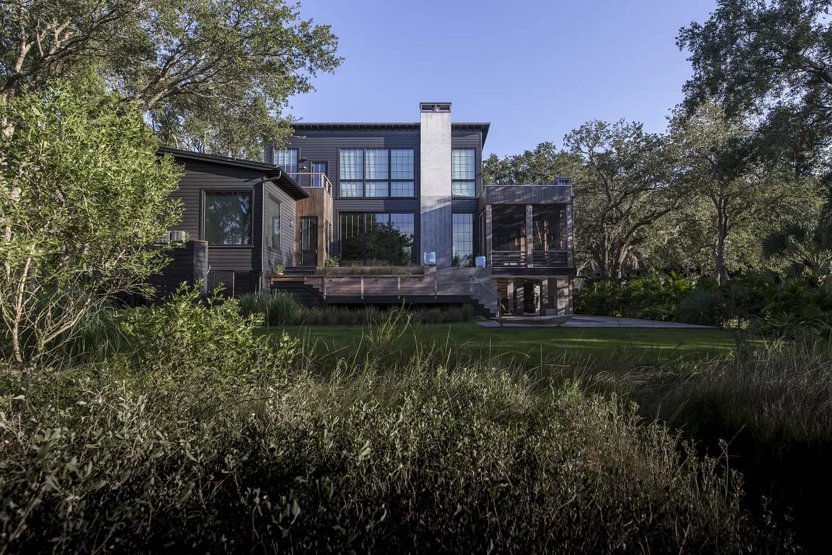 Modern house exterior with large windows, wooden siding, balconies, and surrounding landscaping.