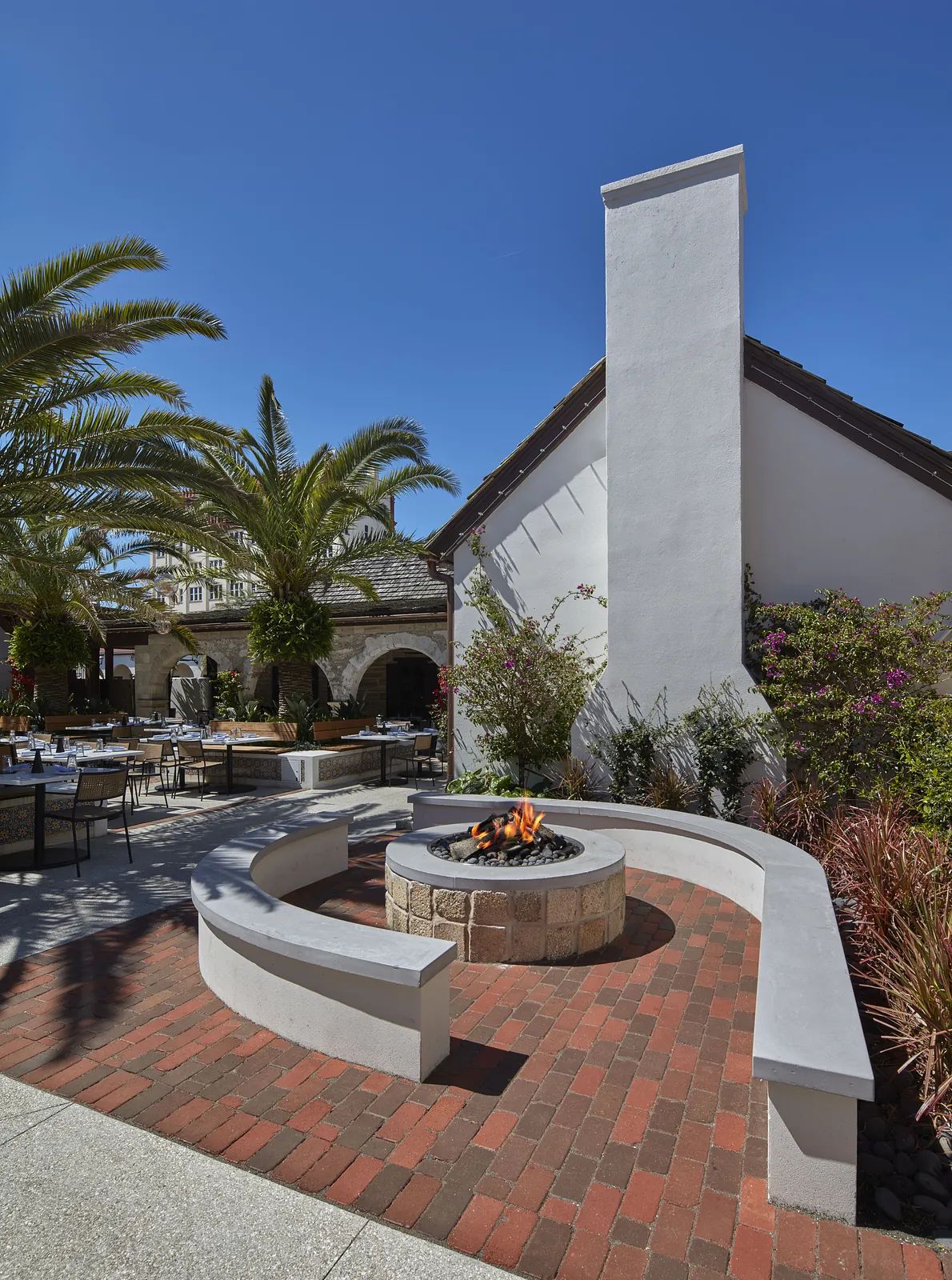 Outdoor patio with circular fire pit, concrete seating, brick flooring, and surrounding greenery.