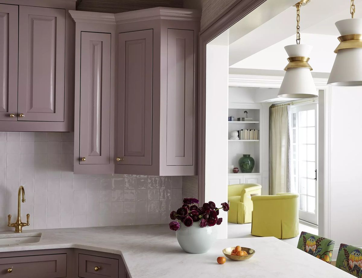 Kitchen with purple cabinetry, marble countertop, gold faucet, and flowers on the counter opening to a bright area.