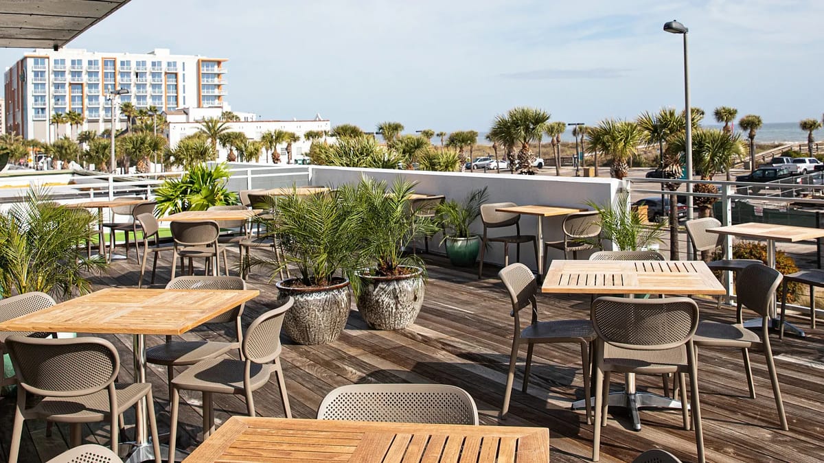 Outdoor patio with wooden tables, gray chairs, large potted plants, and wooden deck overlooking a skyline.