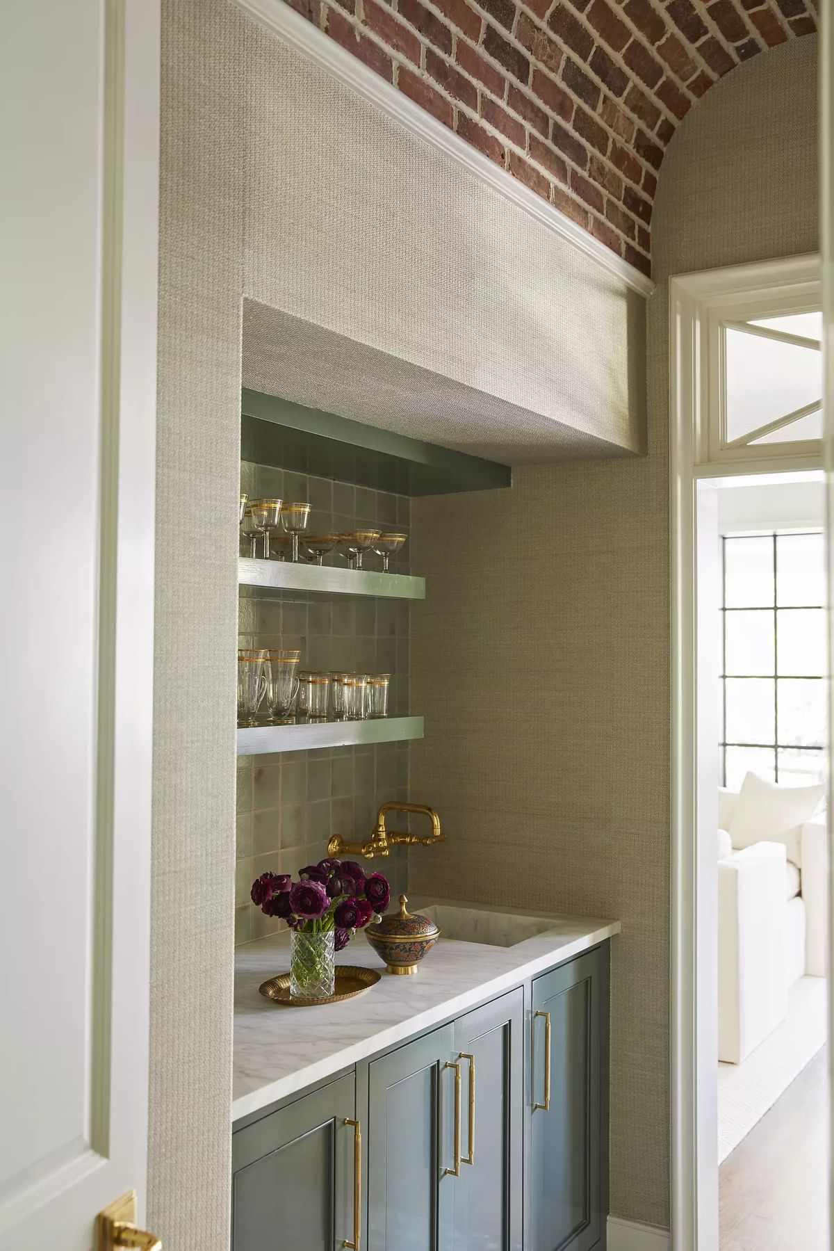 Butler's pantry with blue-green cabinet, marble countertop, glass shelves, and brick archway.