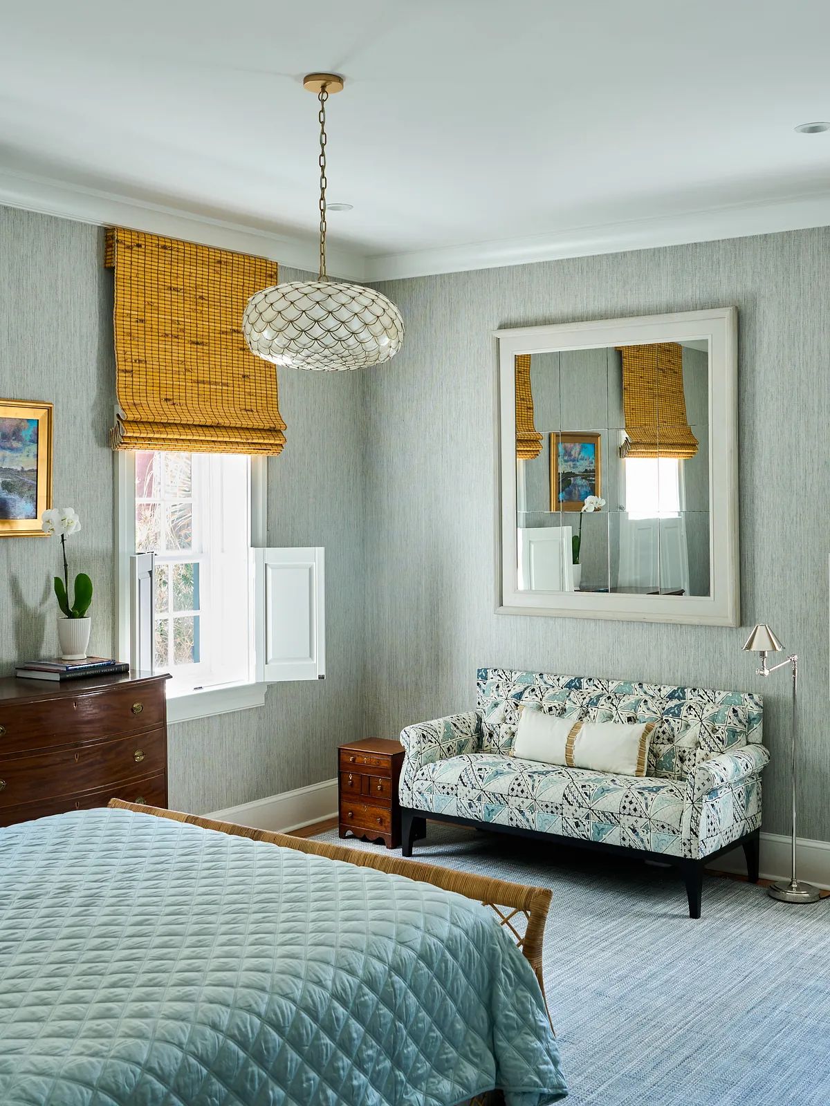 Bedroom with blue quilt, patterned settee, mirror, window with blinds, and wooden dresser.