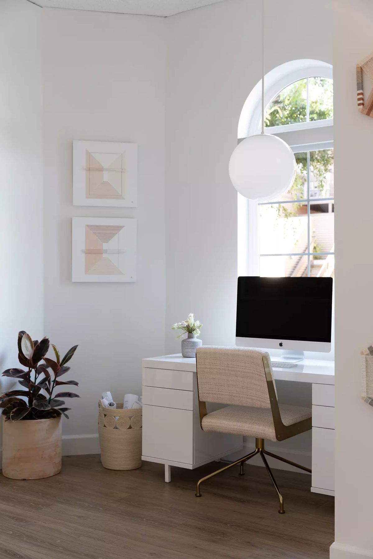 Office with white desk, computer, beige chair, wall art, potted plant, and arched window for natural light