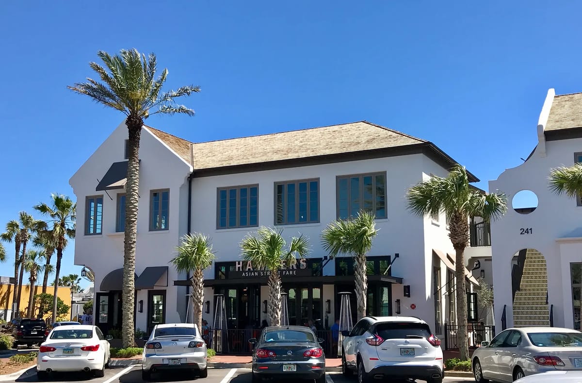 Exterior view of a white building with large windows, palm trees, and a parking area.