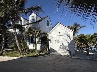 Exterior view of a house with peaked roof, blue shutters, white siding, and palm trees.