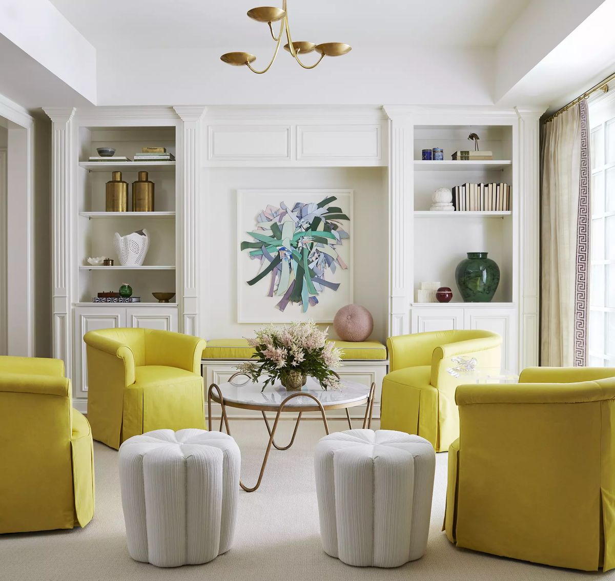 Living room with yellow chairs, glass coffee table, shelves with vases, books, and floral artwork.