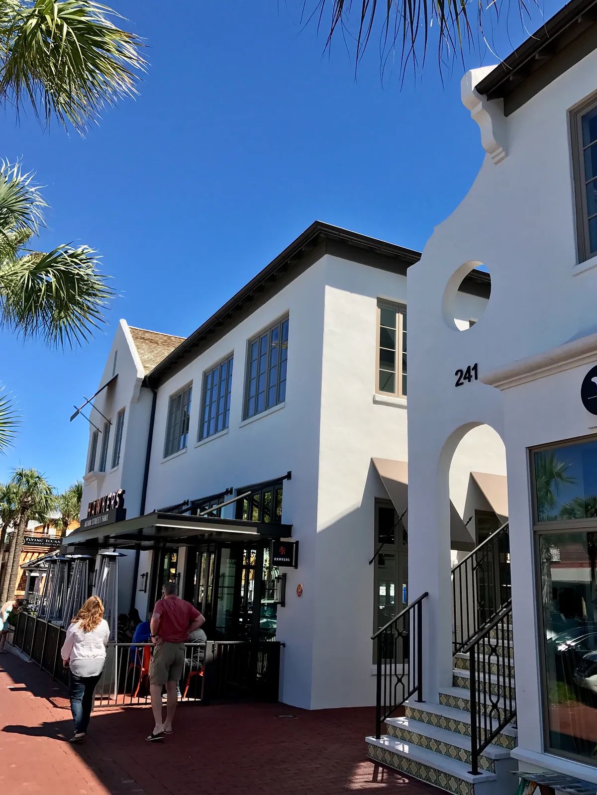 Exterior front of a two-story building with large windows, white stucco, and palm trees.