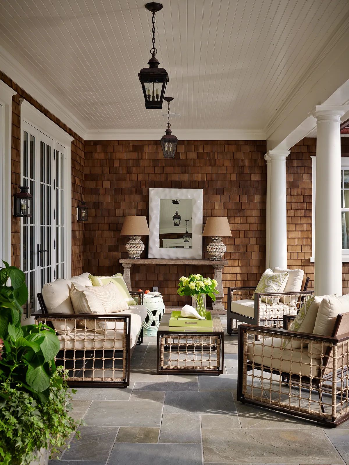 Patio with upholstered chairs, coffee table, potted flowers, wood shingle walls, and stone tile flooring.