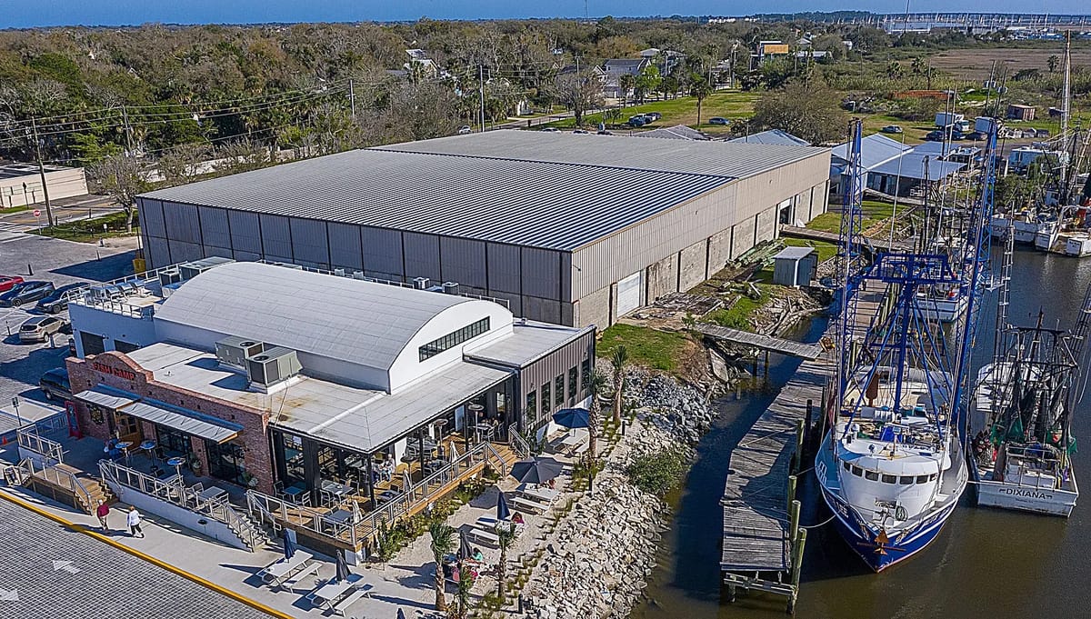 Exterior view of an industrial building and smaller brick structure with outdoor seating and docked boat.