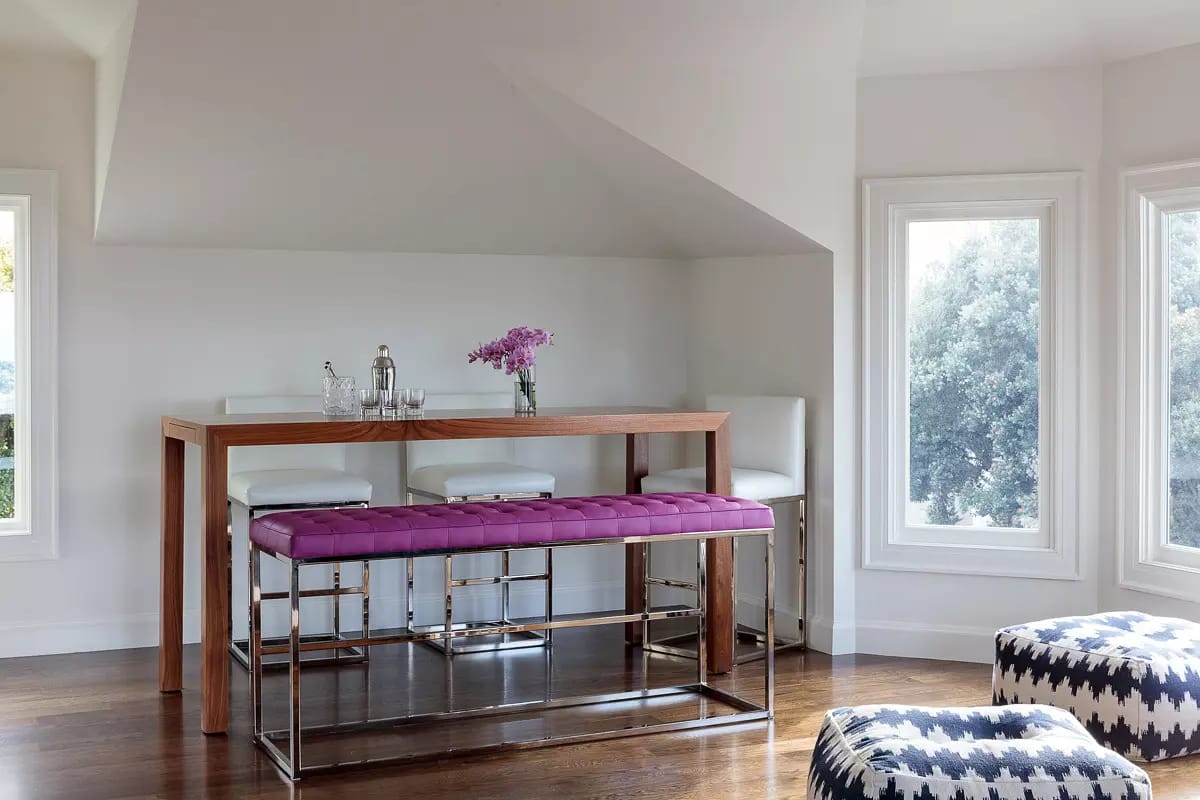 Seating area with wood bar table, white stools, purple bench, and patterned ottoman near large windows.