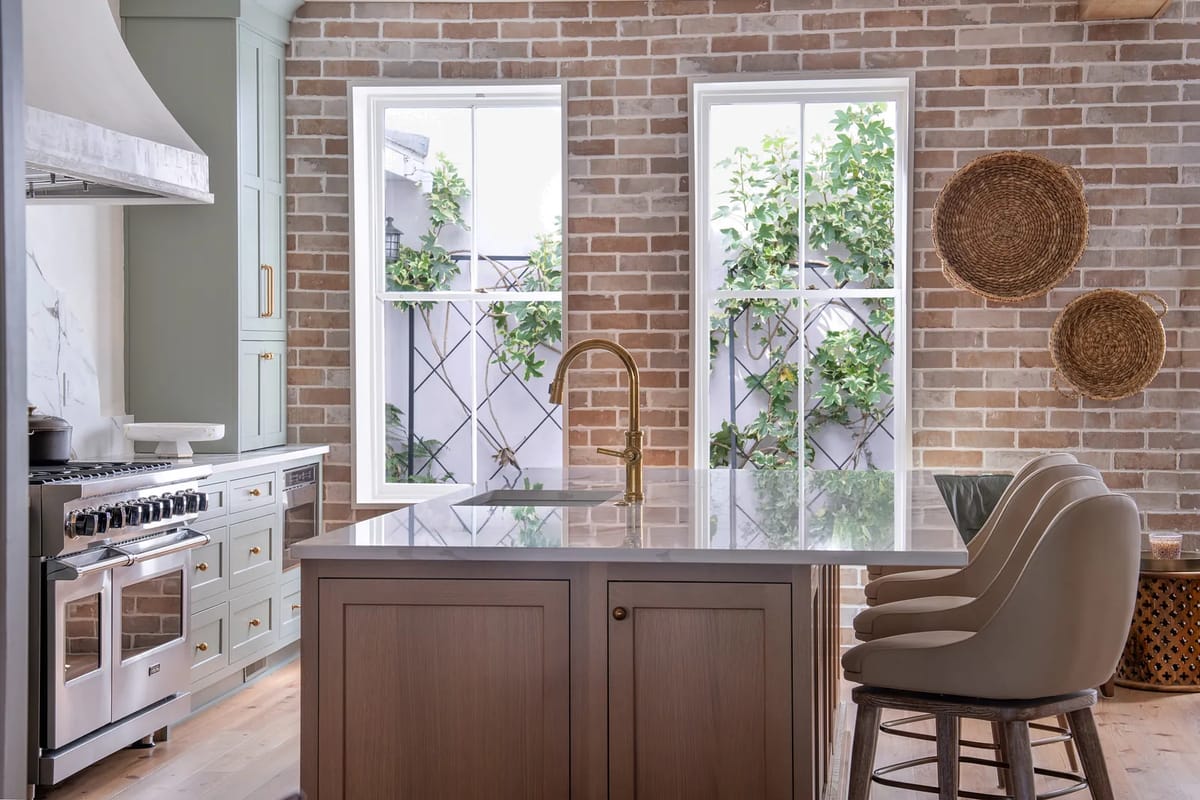 Kitchen with marble island, beige cabinetry, gas range, exposed brick walls, and large windows with vegetation.
