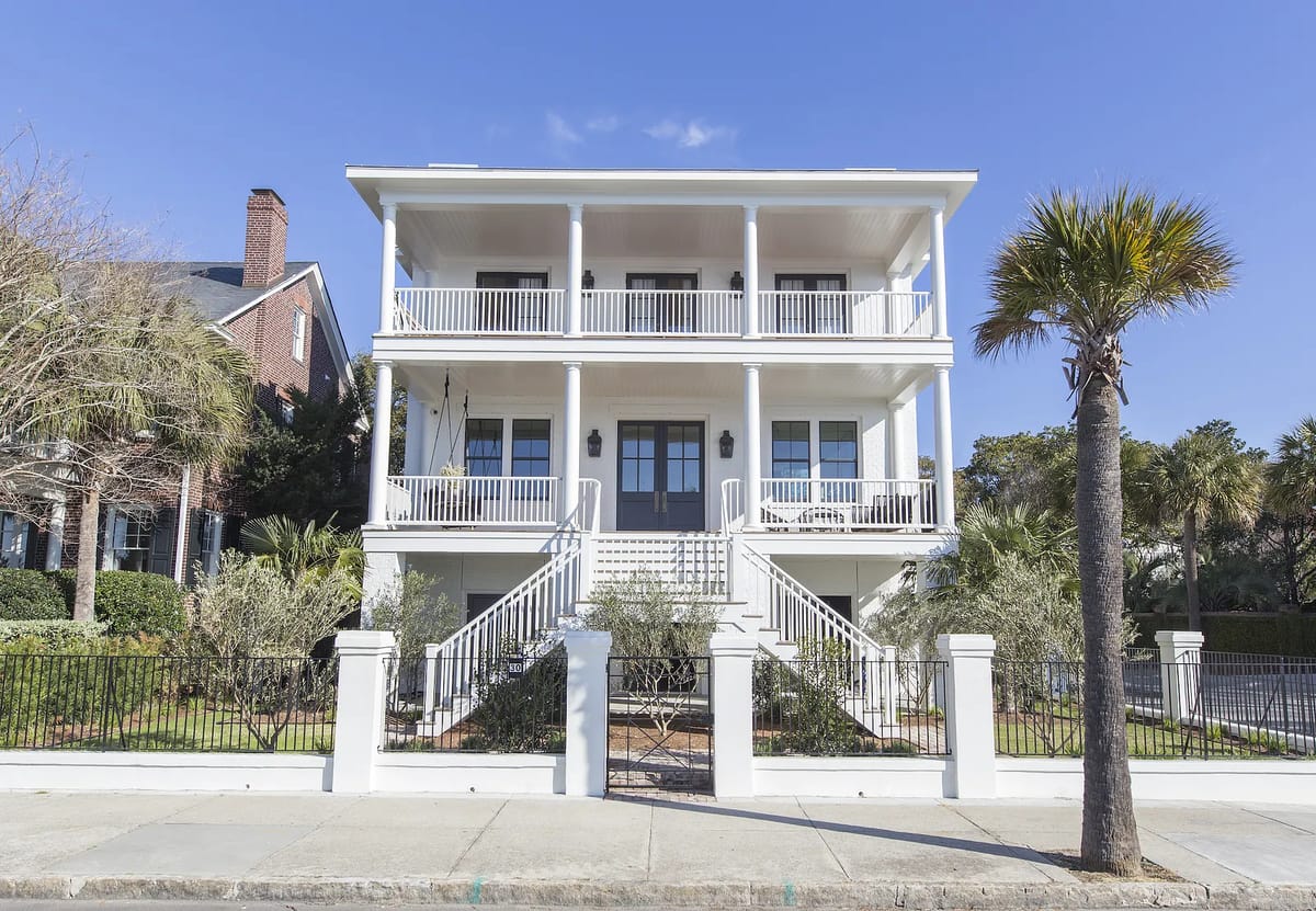 Two-story house exterior with white facade, balconies, staircase, palm trees, and white fence.