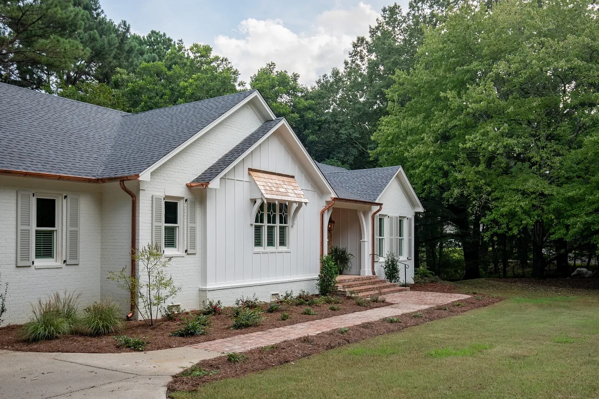 Front exterior of a house with white brick, wooden accents, landscaped shrubs, and a brick pathway.