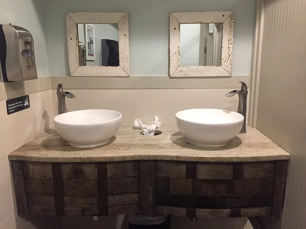 Bathroom with floating wooden vanity, white vessel sinks, rustic mirrors and light-colored tiled walls.