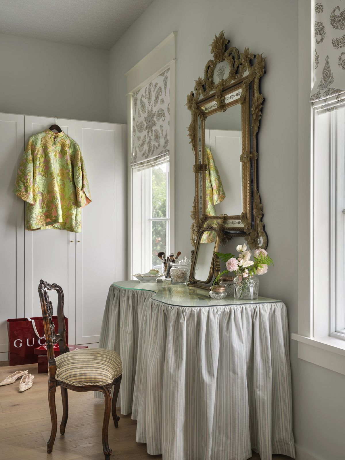 Dressing area with a round table, vintage mirror, striped chair, built-in cabinets, and windows with patterned shades.